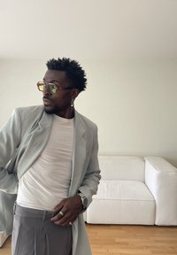 Muž v sivom saku, bielom tričku, sivých nohaviciach, žltých slnečných okuliaroch a perlovej náušnici pózuje v interiéri vedľa bielej pohovky.