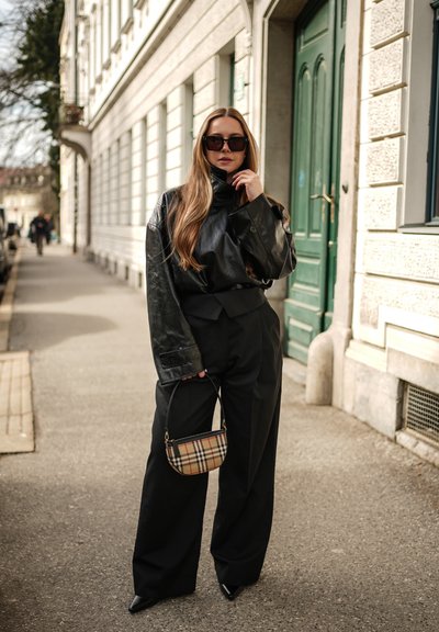Mujer con cabello largo que lleva una chaqueta de cuero negra, pantalones anchos negros, gafas de sol grandes y sostiene un pequeño bolso de mano a cuadros en la acera de una ciudad.