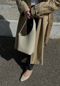 Sac en cuir beige porté à l'épaule avec une texture lisse, un design courbé, associé à un pantalon noir et des ballerines nude ; un trench-coat beige complète le look.