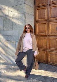 Brązowa zamszowa kurtka, jasnoróżowy sweter, ciemnoszare szerokie spodnie i brązowe sneakersy. Trzyma małą brązową torbę, stojąc obok drewnianych drzwi.