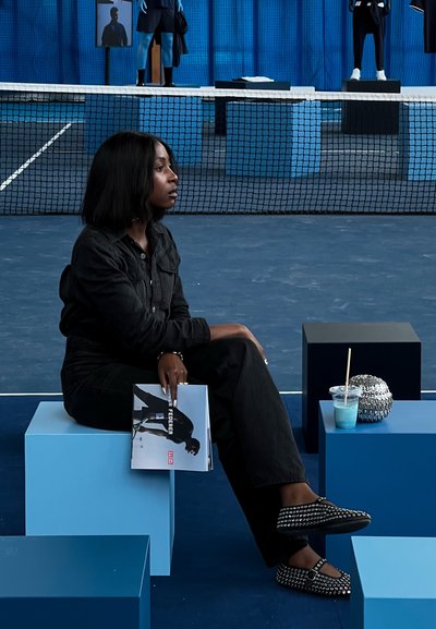 Una persona vestida de negro está sentada sobre un cubo azul, sosteniendo una revista. Cerca, se puede ver un objeto texturizado de color plateado y una bebida con una pajita.