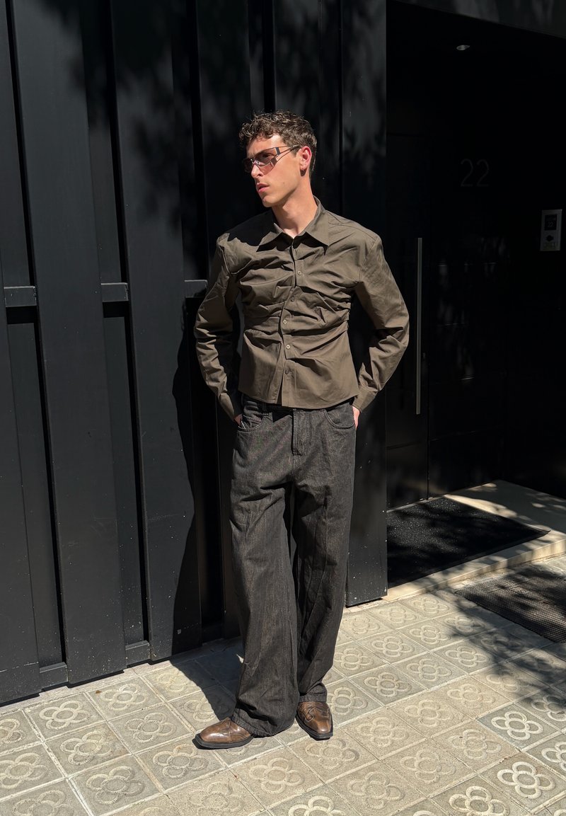 Un homme portant des lunettes de soleil, une chemise olive ajustée, un pantalon large foncé et des chaussures brunes s'adosse à un mur noir sous une forte lumière du soleil.