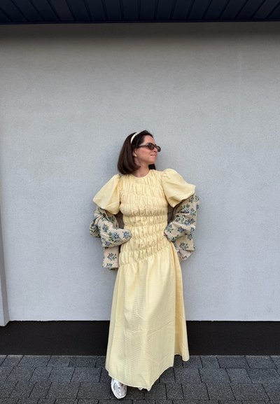 Mujer vestida con un largo vestido amarillo de mangas abullonadas y una chaqueta floral colgada sobre los brazos, de pie frente a una pared lisa de color gris claro.
