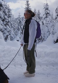 A person standing in the snow, dressed in a black puffer vest, a grey sweatshirt, green trousers, and white trainers, holding a lead attached to a dog. They have a purple backpack.