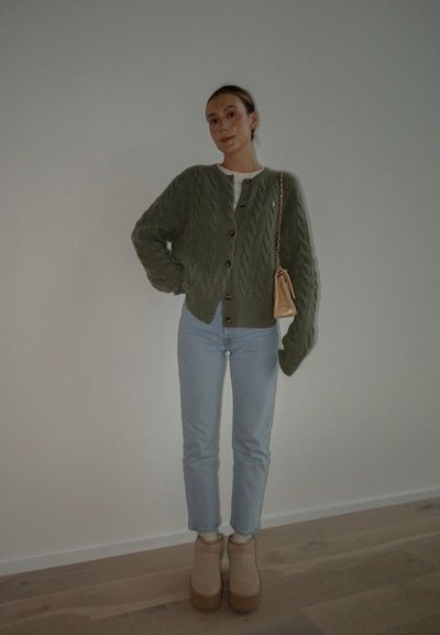 Cárdigan verde de punto trenzado con botones oscuros, camisa blanca debajo, jeans azul claro y botas de tobillo de plataforma beige. Bolso beige con cadena.