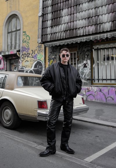 Chaqueta bomber negra, suéter negro y pantalones de cuero negro brillantes. Gafas de sol rectangulares plateadas. Coche vintage de color claro de fondo.