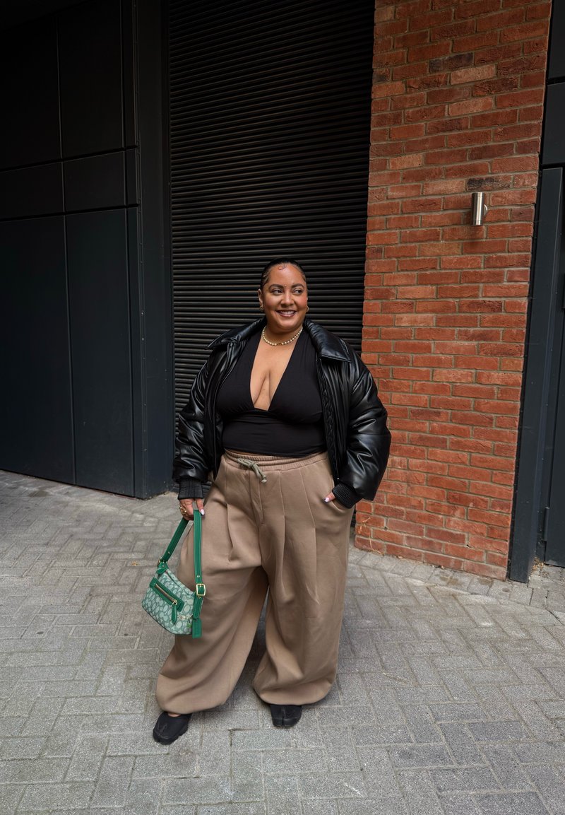 Black puffer jacket, black fitted top, wide beige trousers, green handbag with patterns, and black shoes. Background features brick wall and black shutter.