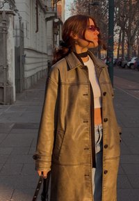 Καφέ ψεύτικο δερμάτινο παλτό με αντίθετη ραφή, κουμπιά και πλαϊνές τσέπες, φορεμένο πάνω από λευκό crop top και μπλε τζιν παντελόνι.