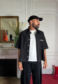 Μαύρο κοντομάνικο πουκάμισο με λευκή ραφή και μπροστινή τσέπη, φορεμένο πάνω από λευκό μπλουζάκι, συνδυασμένο με μαύρο παντελόνι με φαρδιά γραμμή.