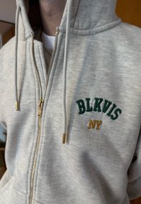 Personne portant un sweat à capuche gris clair à fermeture éclair avec "BLKVIS NY" brodé en vert et or sur la poitrine, capuche tirée sur la tête.