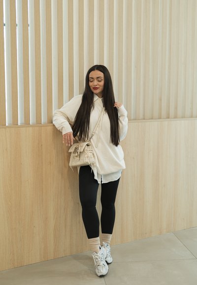 Mujer con cabello largo y oscuro, vestida con suéter blanco, leggings negros, calcetines beige y zapatillas, apoyada contra una pared de paneles de madera, sosteniendo un bolso beige.