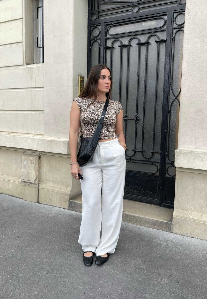 Μπεζ τοπ με σχέδιο ζώου, άσπρο παντελόνι σε φαρδιά γραμμή, μαύρη τσάντα ώμου και μαύρες φλατ εσπαντρίγιες; στέκεται κοντά σε διακοσμητική πόρτα.