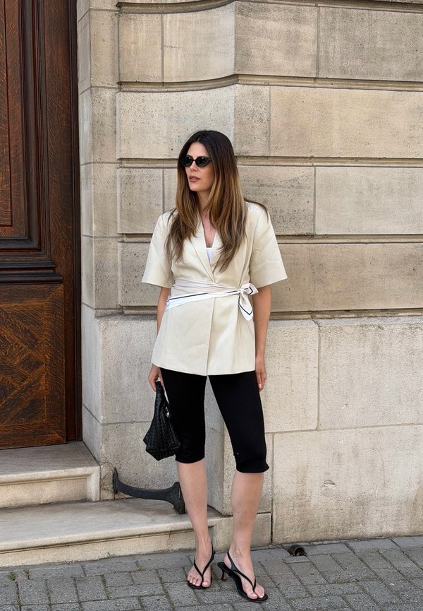 Kvinde med langt hår iført solbriller, beige wrap-blazer, sorte korte leggings, sorte hælede sandaler, holder en sort flettet taske, står ved en stenmur.
