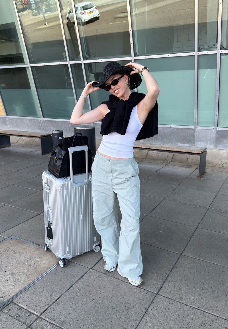 Femme ajustant un chapeau de soleil noir, portant un débardeur blanc, un pantalon clair, des lunettes de soleil, debout sur un trottoir avec une valise argentée et un sac noir près d'un bâtiment en verre.