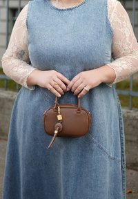 Farmerruha kék árnyalattal és hosszú csipkés ujjakkal. Egy kis barna bőr kézitáskát tart arany díszítésekkel és kerek fogantyúval.