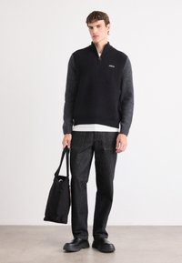 Zwart en grijs kleurblokken gebreide trui met een half ritskraag, gecombineerd met donkere jeans en een zwarte schoudertas. Soepele textuur en eenvoudig ontwerp.