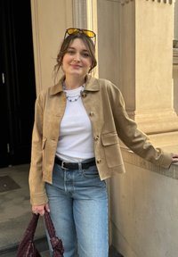 Beige semsket jakke med krage, frontknapper og lomme. Stylet med en hvit t-skjorte, blå jeans og en mørk vevd håndveske.