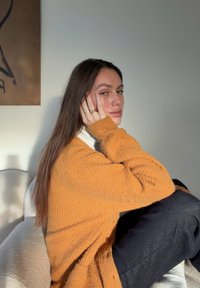 Narancssárga, bordázott textúrájú kötött kardigán fehér garbó fölött, világos háttér előtt; tompa fekete nadrág és laza póz.