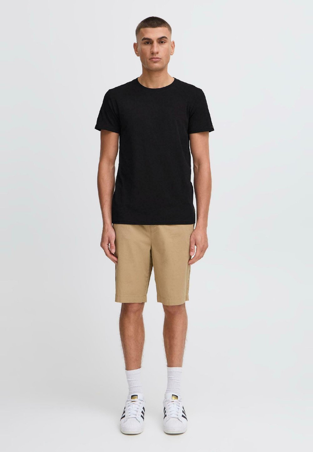 T-shirt nera in cotone a maniche corte abbinata a pantaloni corti beige. Il modello indossa sneakers bianche e calze bianche, in piedi su uno sfondo semplice.