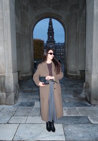 Brauner Übermantel, grauer Rock und schwarze Stiefel. Stehend in einem Durchgang, eine schwarze Clutch haltend. Steinboden und ein historisches Gebäude im Hintergrund.