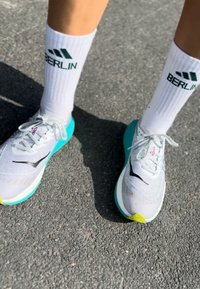 Könnyű futócipők fehér hálós felsőrésszel, fekete díszítésekkel és tengerkék talppal. Fehér zoknival párosítva, amelyen zöld "BERLIN" felirat található.