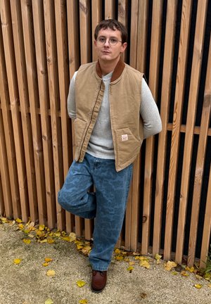 Chaleco de algodón color canela sobre un suéter gris, combinado con jeans azules con estampado y zapatos marrones. Listones de madera en el fondo.