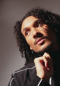 Μαύρο μπουφάν με λευκή ρίγα, σγουρά μαλλιά, ανοιχτόχρωμο δέρμα, σκουλαρίκια, χέρι resting στο πηγούνι, απαλή φωτισμός δημιουργεί σκιές στο πρόσωπο.