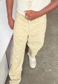 Beige Baumwollhosen mit einer lockeren Passform, ausgestattet mit Fronttaschen und einem Kordelzugbund, kombiniert mit weißen Sneakers mit roten Akzenten.