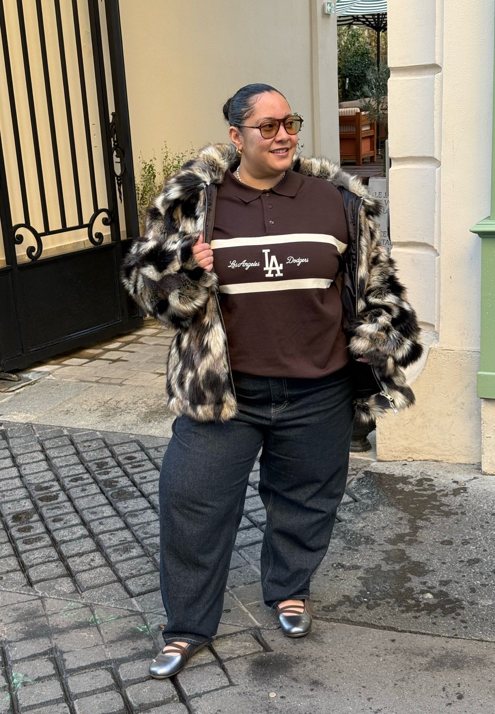 Manteau en fausse fourrure avec des motifs gris et noirs, polo marron avec logo blanc LA, jean foncé, et ballerines argentées sur une rue pavée.