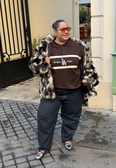 Abrigo de piel sintética con patrones grises y negros, polo marrón con logo blanco de LA, jeans oscuros y zapatos plateados planos sobre calle empedrada.