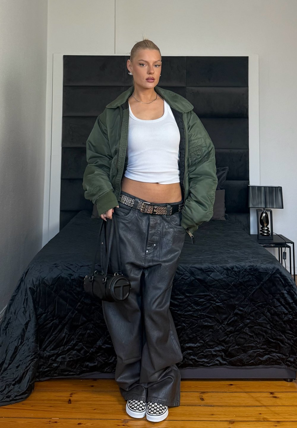 Kobieta stoi w sypialni, ubrana w zieloną kurtkę, biały top na ramiączkach, czarne skórzane spodnie, buty w kratkę, trzyma czarną torebkę.