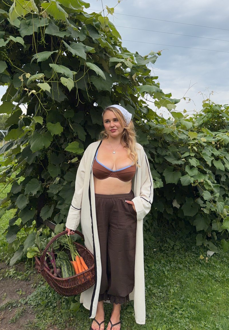 Femme debout à l'extérieur près de plantes feuillues, portant un haut et un pantalon marron avec une robe légère, tenant un panier de légumes frais comprenant des carottes et des concombres.