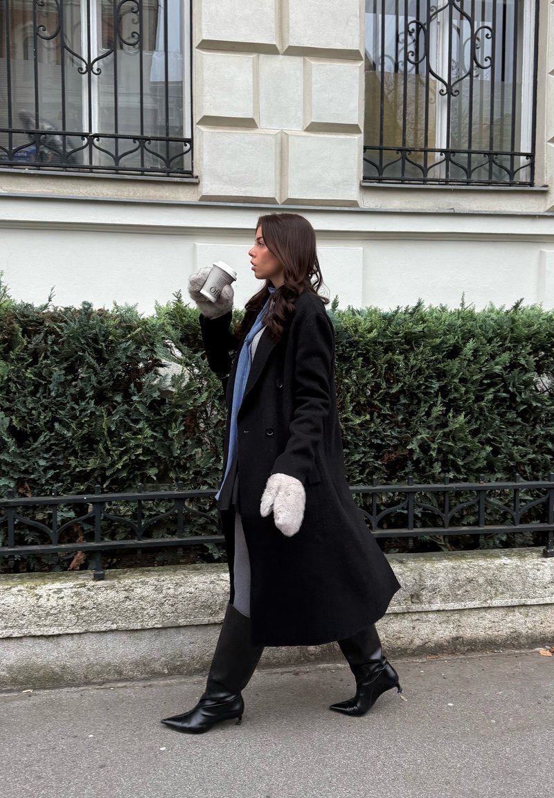 Manteau noir croisé, mitaines grises, chemise bleue, jupe grise et bottes noires montant aux genoux. Tenant une tasse de café blanche, marchant près de la verdure.