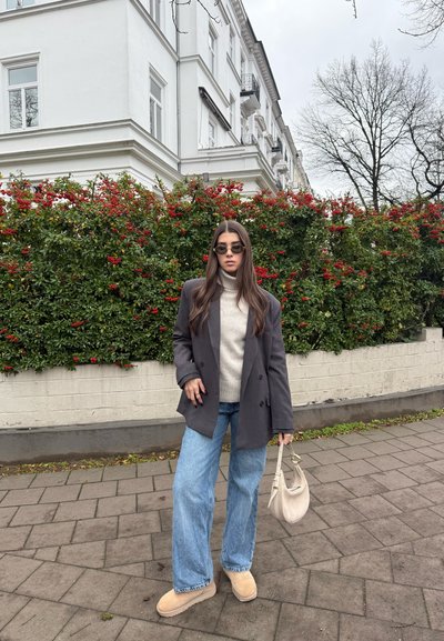 Joven mujer con gafas de sol, blazer gris, suéter beige, vaqueros azules y zapatos marrones parada en la acera con arbustos de bayas rojas y un edificio blanco detrás.