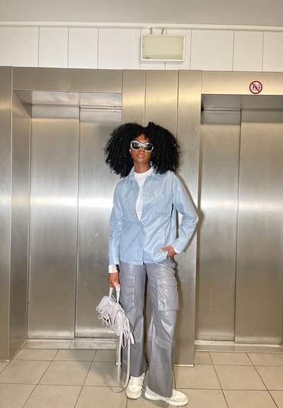 Chaqueta de cuero azul claro, camisa blanca, pantalones gris de pierna ancha, bolso plateado y zapatillas blancas. Cabello rizado negro, grandes gafas de sol. Fondo de ascensor metálico.