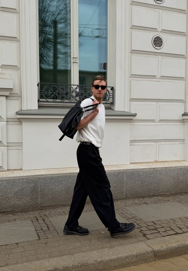 Homme portant des lunettes de soleil, une chemise blanche et un pantalon noir marchant sur le trottoir avec un sac à dos noir porté sur l'épaule à côté d'un bâtiment.