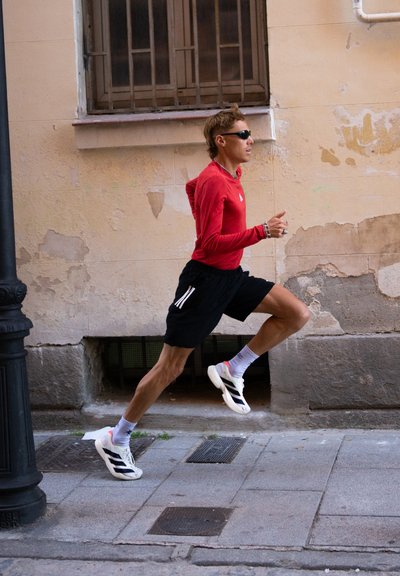 Una persona corre por una calle, vistiendo una camiseta de manga larga roja, pantalones cortos negros y zapatillas deportivas blancas con rayas negras.