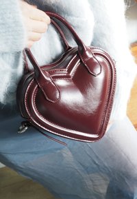 Pessoa vestindo um suéter felpudo azul claro segura uma bolsa de mão em forma de coração, de couro vermelho escuro, pelas alças.