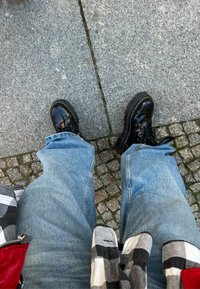 Černé lakované kozačky se šněrováním, s žlutým prošitím, spárované se světle modrými džíny. Pozadí tvoří šedá kostková dláždění.