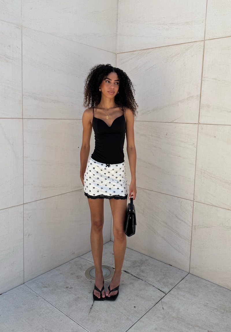 Jeune femme aux cheveux bouclés portant un haut noir et une mini-jupe blanche avec des détails noirs, tenant un sac à main noir, debout contre un mur carrelé.