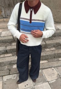 Osoba w białym, puszystym swetrze w niebieskie i brązowe paski, ciemnych dżinsach o szerokich nogawkach, brązowych butach, pierścionkach oraz czarnej torbie na ramię na świeżym powietrzu.