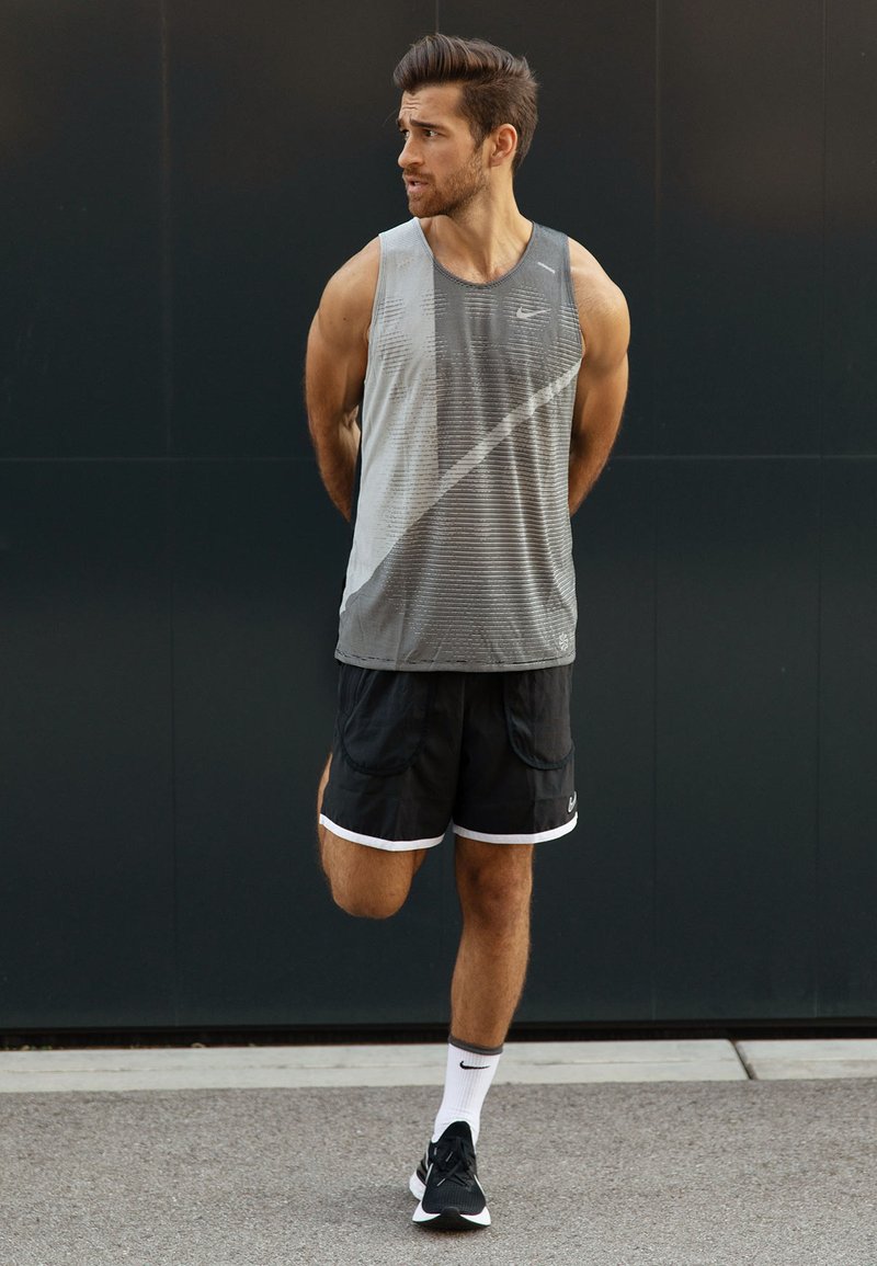 Grijze sportieve tanktop met horizontale strepen, zwarte shorts met witte accenten en zwarte hardloopschoenen. Model poseert op een grijze ondergrond.
