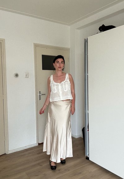 Mujer de pie en interiores con un top blanco de encaje sin mangas, una falda larga de satén color crema, zapatos negros y pendientes de aro plateados.
