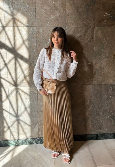 Joven mujer de pie contra una pared de mármol, vistiendo una blusa de encaje blanco, falda dorada plisada, sandalias blancas y sosteniendo un bolso de mano floral.