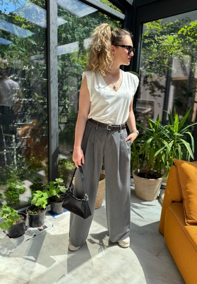 Pantalones de pierna ancha en gris claro, top sin mangas blanco, bolso negro y zapatillas claras. Entorno interior con vegetación y un sofá amarillo.