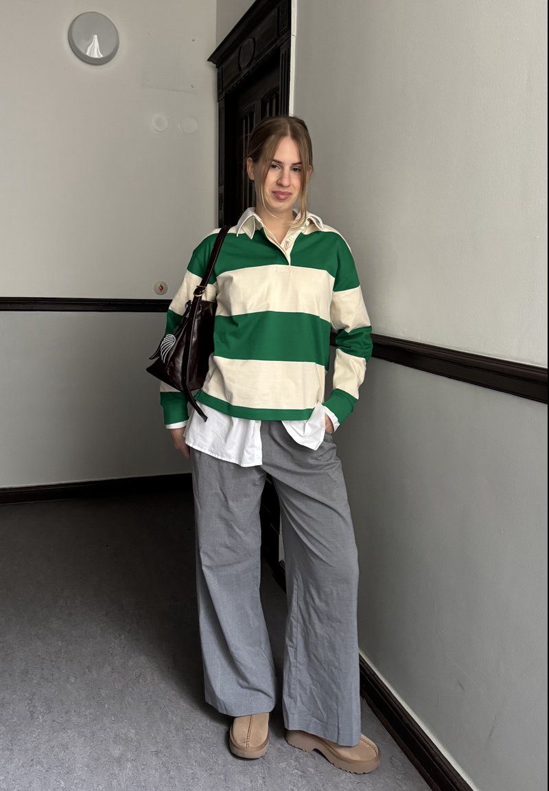 Grün- und cremefarben gestreiftes Rugbyshirt über einem weißen Hemd mit Knöpfen, kombiniert mit grauen Hosen mit weitem Bein und beigen Schuhen, dazu eine dunkle Tasche.