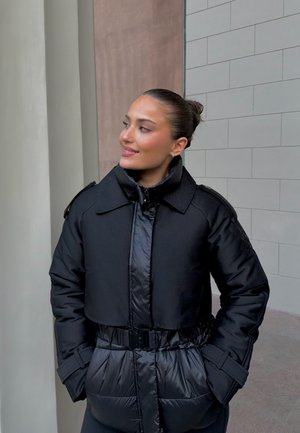 Chaqueta acolchada negra con diseño corto, cintura con cinturón y texturas contrastantes. Cierre de cremallera y detalles en los hombros visibles.