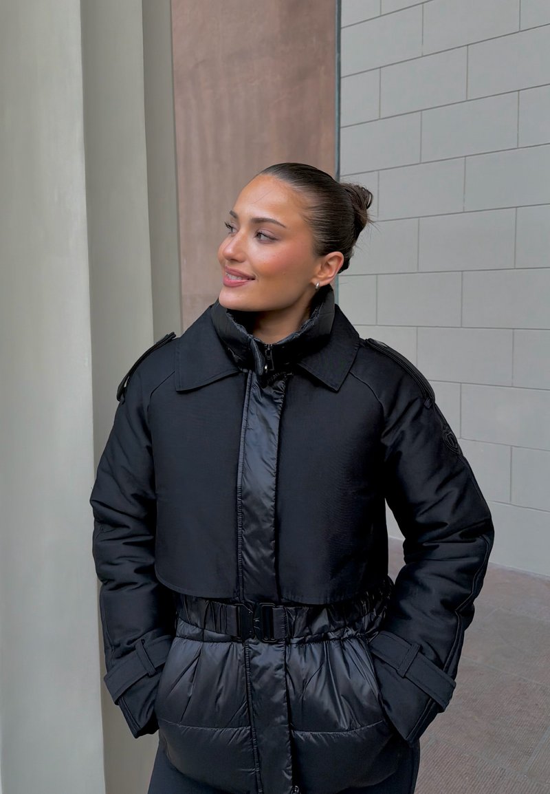 Doudoune noire présentant un design court, une taille cintrée avec ceinture, et des textures contrastantes. Fermeture éclair et détails aux épaules visibles.