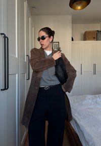 Manteau en daim marron, pull gris, pantalon noir à jambes larges et lunettes de soleil noires. Posé dans une pièce lumineuse avec une literie blanche et des rangements.
