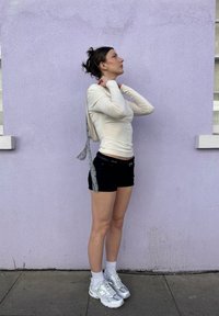 Un top crem cu mâneci lungi și un bretele cu buline gri, pantaloni scurți negri și șosete albe până la gleznă, asortate cu adidași gri. Pe un fundal de perete pastel violet.